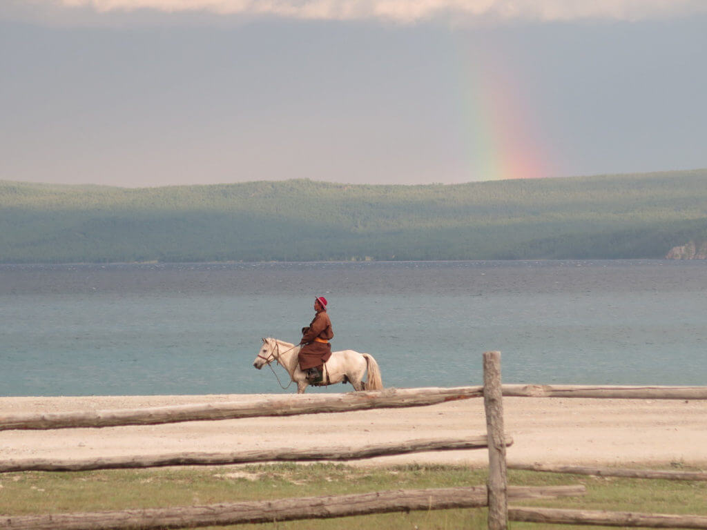 Mann auf weißem Pferd am Chuwsgulsee, im Hintergrund dunkle Wolken und Regenbogen