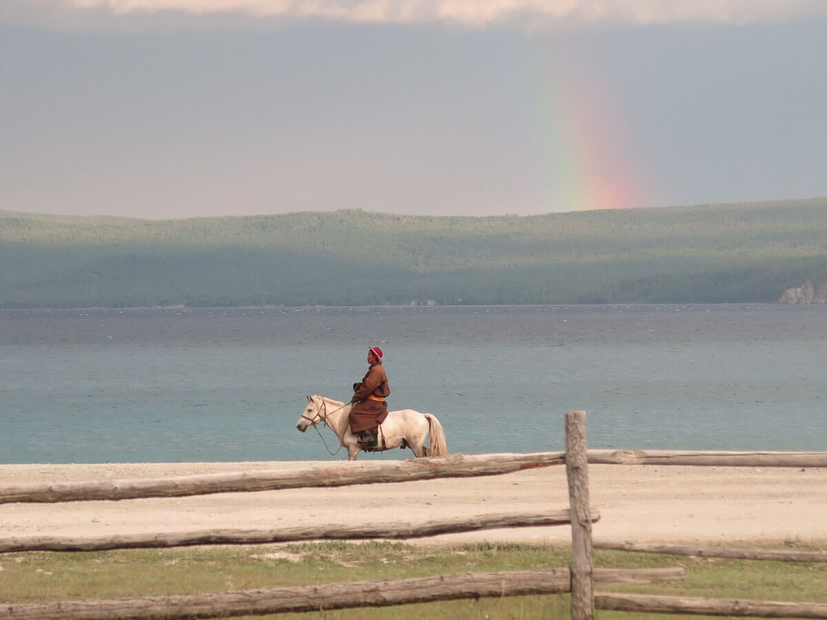 Mann auf weißem Pferd am Chuwsgulsee, im Hintergrund dunkle Wolken und Regenbogen