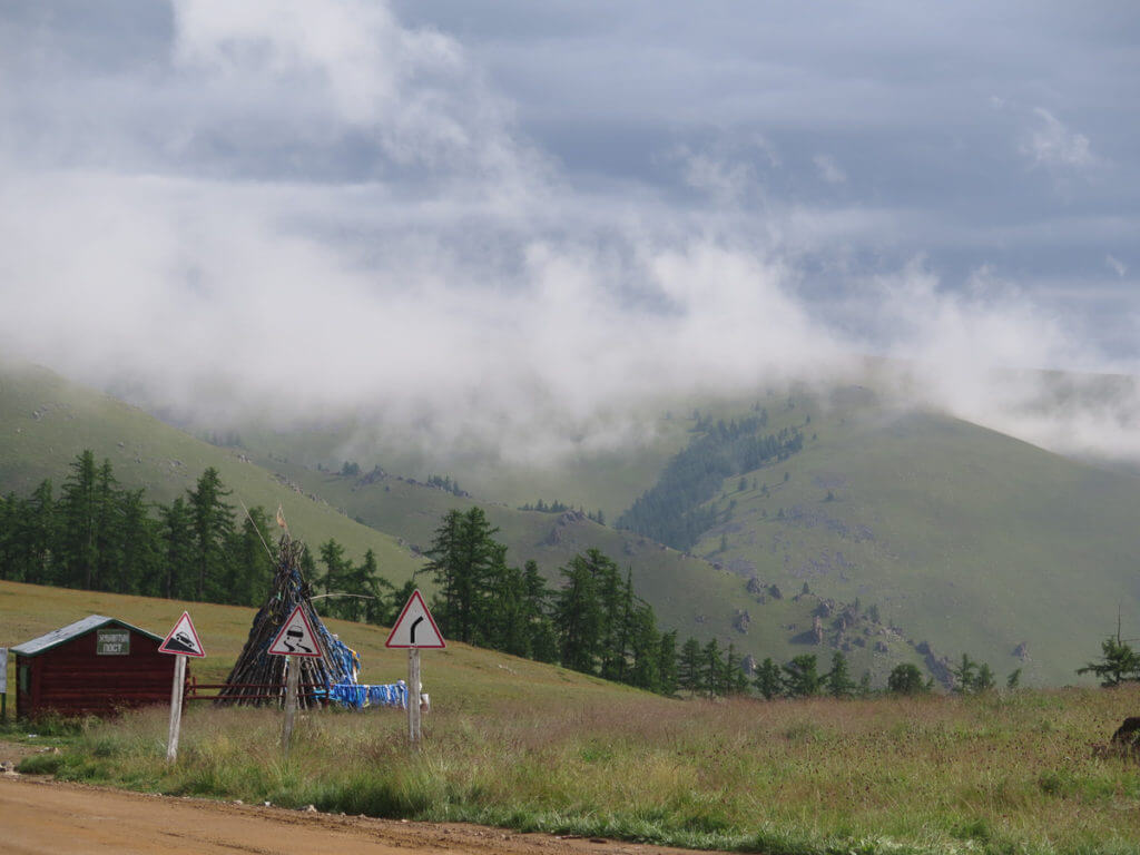 Grüne Berge in der Mongolei mit Wolken und im Vordergrund Warnschilder und ein Owoo