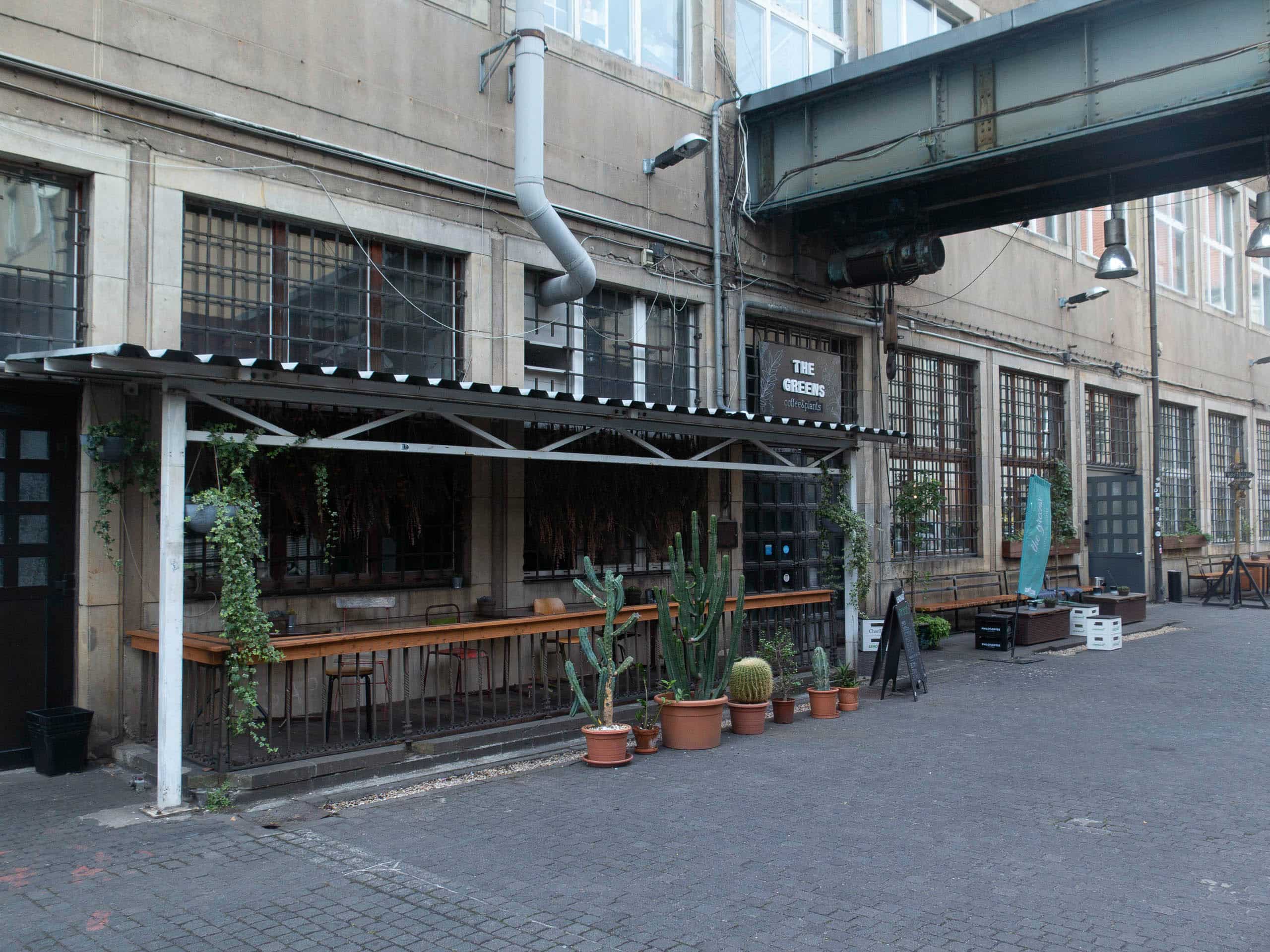 Außenansicht Cafe The Greens, überdachte Terrasse davor mit Stühle, vor der Terasse Pflanzen, industrial
